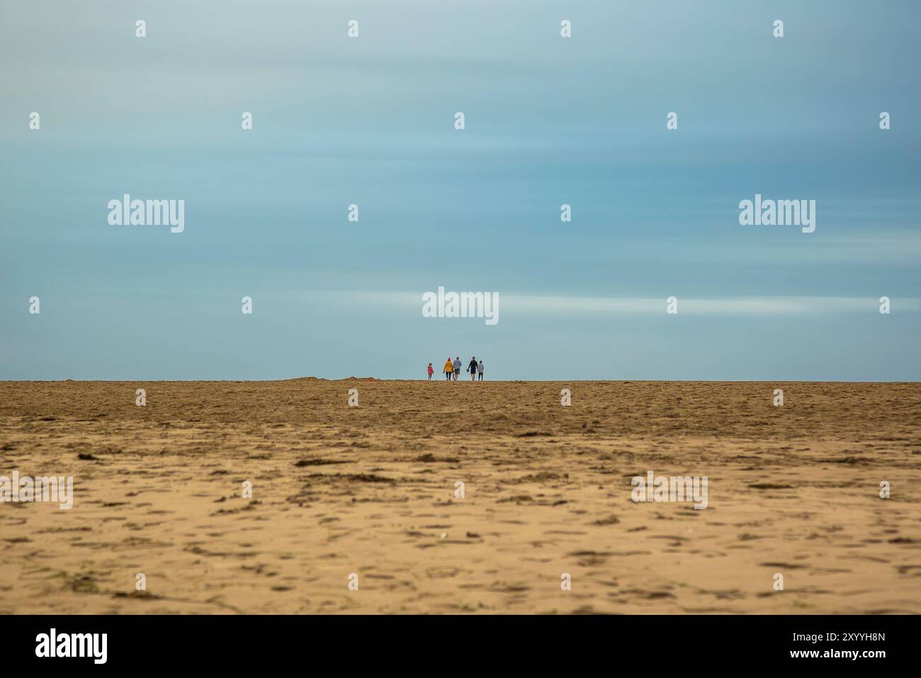 Vista posteriore di una famiglia isolata insieme, in prima serata, a piedi in lontananza, sulla sabbia di una spiaggia del Regno Unito adagiata su un cielo blu. Foto Stock