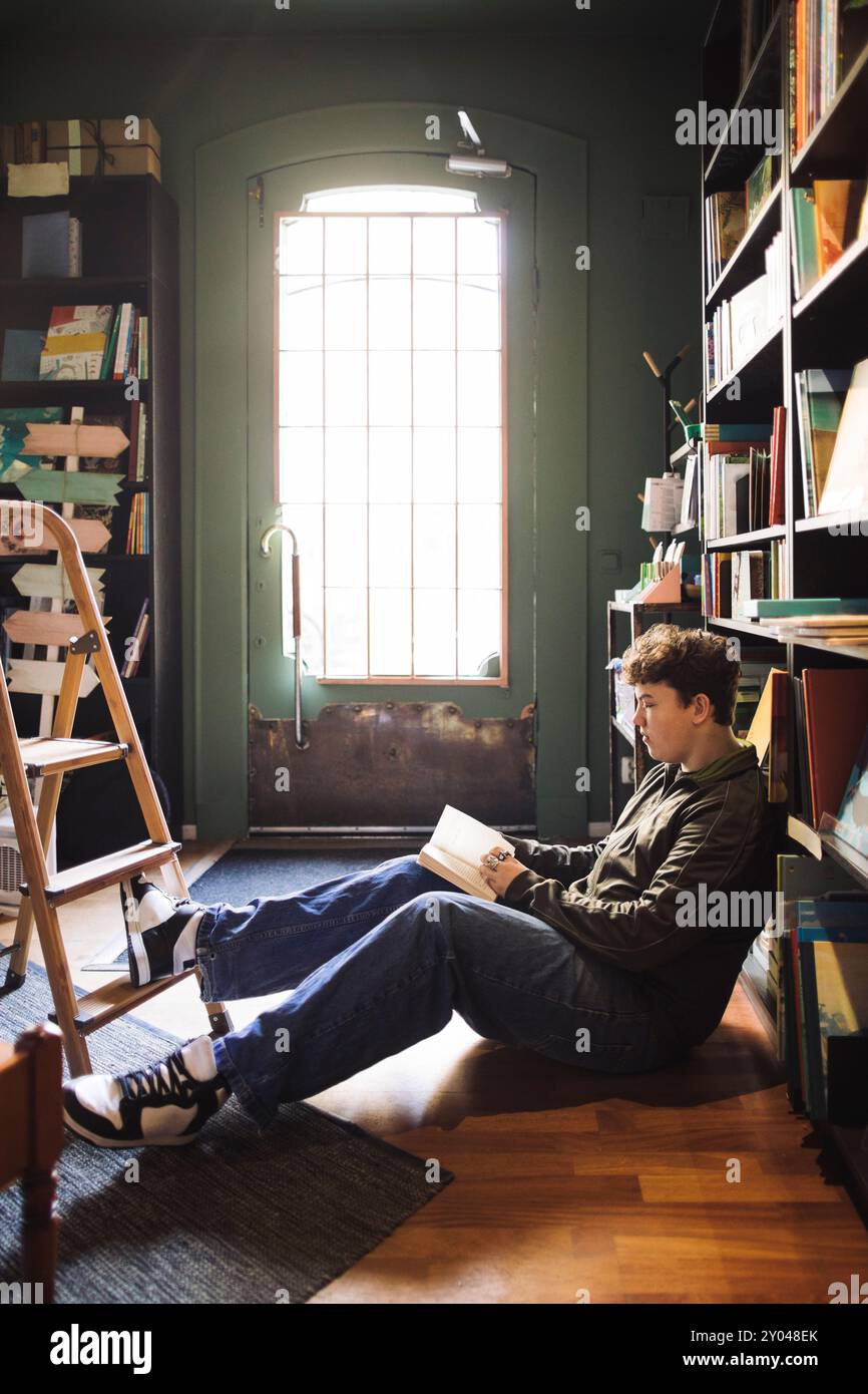 Vista laterale dell'uomo che legge un libro seduto sul pavimento vicino allo scaffale della libreria Foto Stock