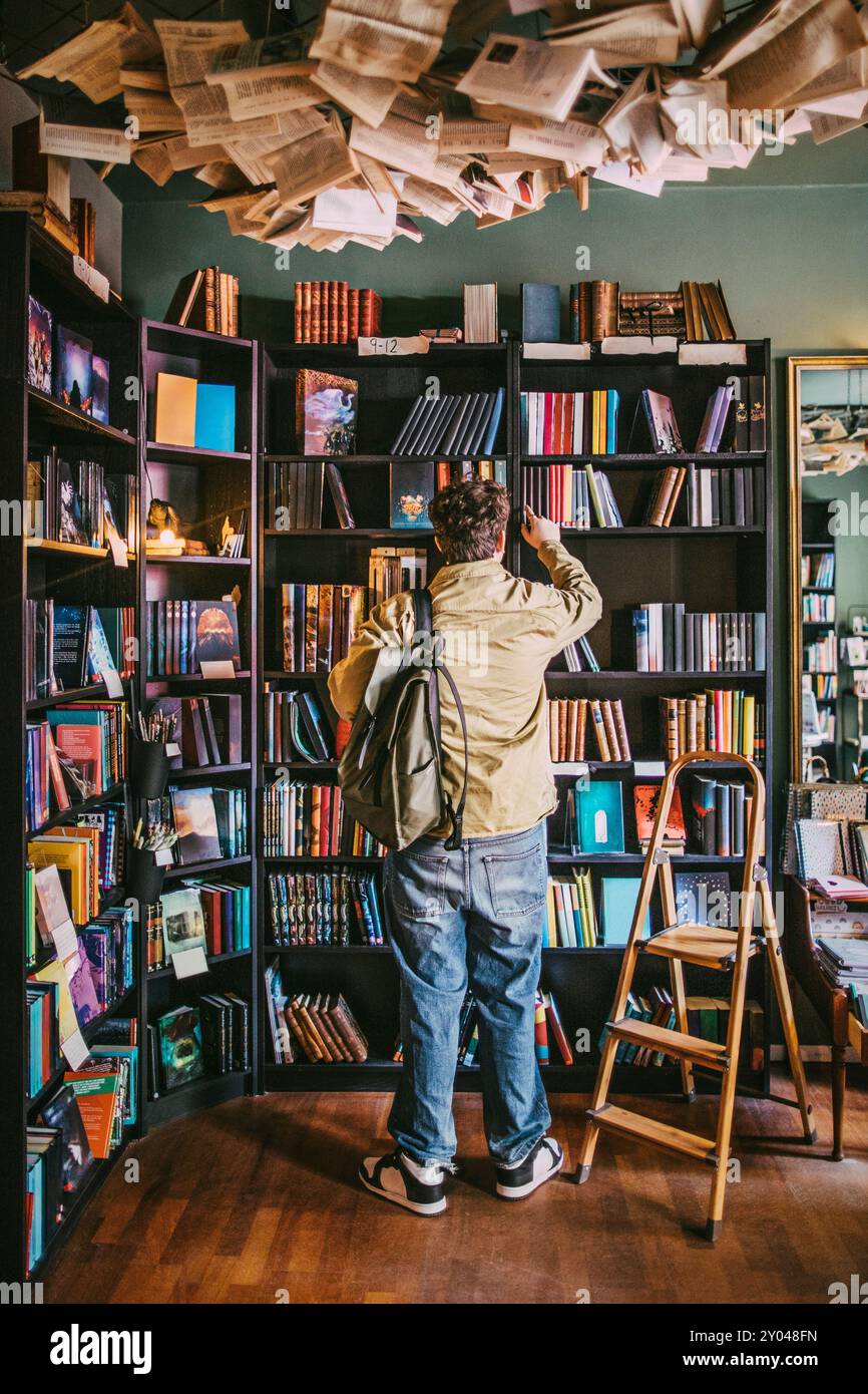 Vista posteriore del giovane con zaino che sceglie libri dallo scaffale della libreria Foto Stock