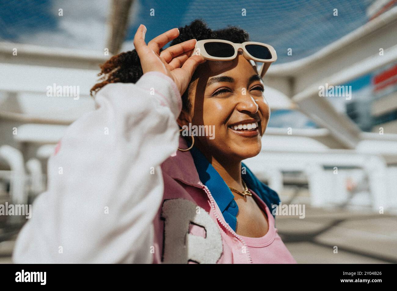 Felice donna alla moda capelli ricci nella giacca letterman con occhiali da sole nel parcheggio Foto Stock