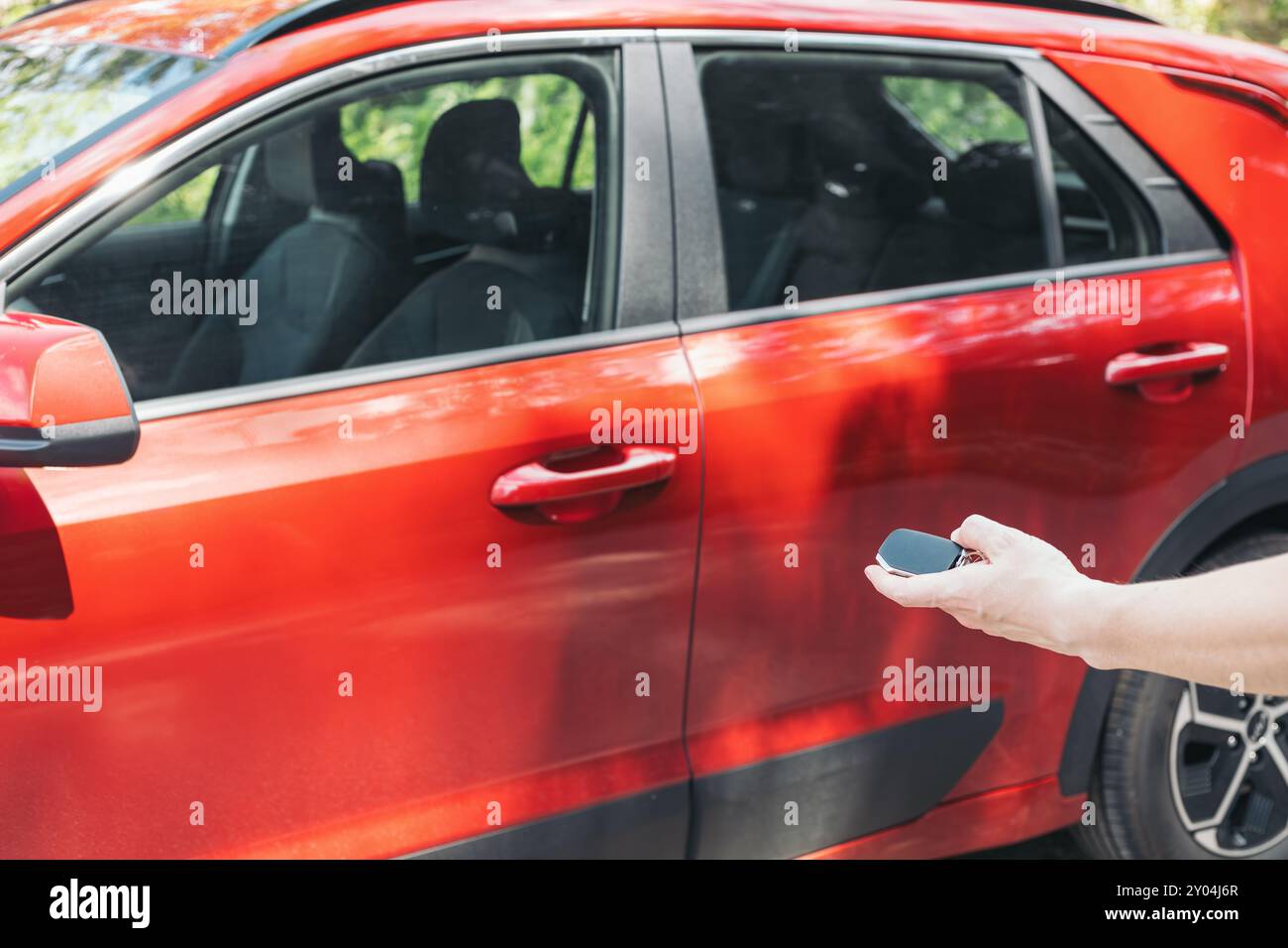 Primo piano di una mano che tiene una chiave accanto a un veicolo rosso, concetto di accesso e controllo remoto dell'auto. Momenti quotidiani associati al proprietario dell'auto Foto Stock