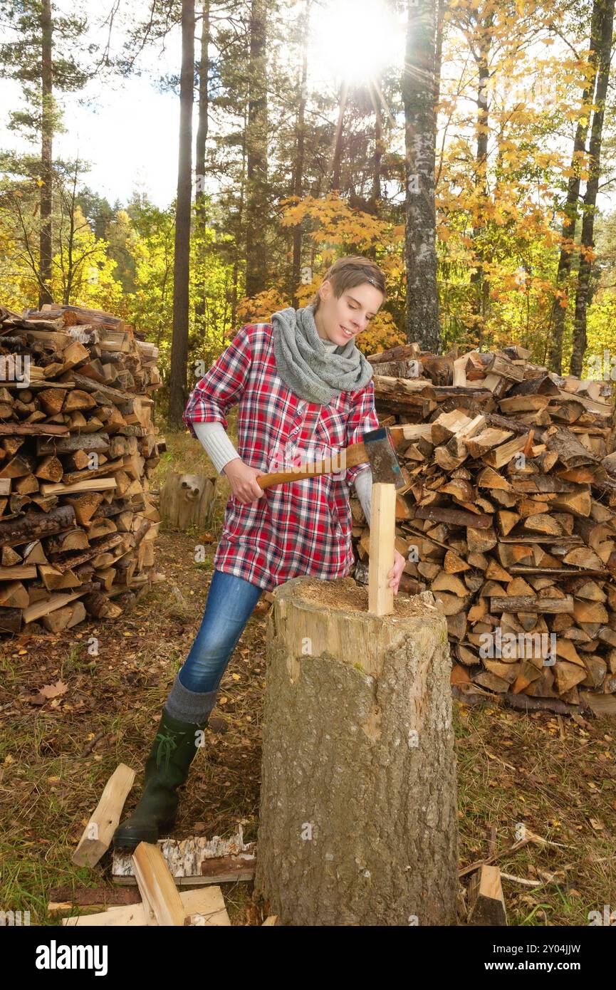 Giovane donna attraente che taglia e divide i tronchi con un'ascia su un blocco di triturazione. La donna indossa abiti da lavoro o per il tempo libero (stivali, jeans, tacca rossa Foto Stock