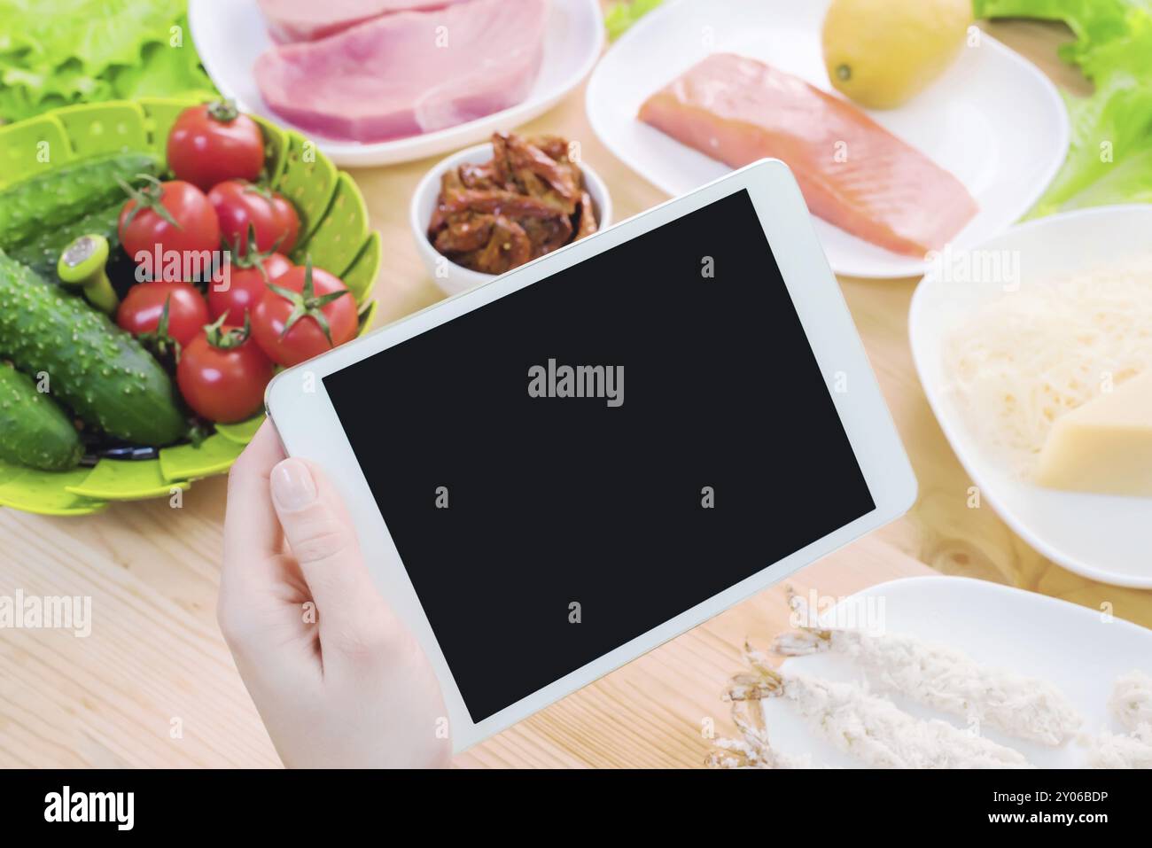 Vista dall'alto ravvicinata di mani femminili che tengono in mano un tablet computer con uno schermo vuoto circondato da cibo sano su un tavolo da cucina di casa. Pezzo da infilare Foto Stock