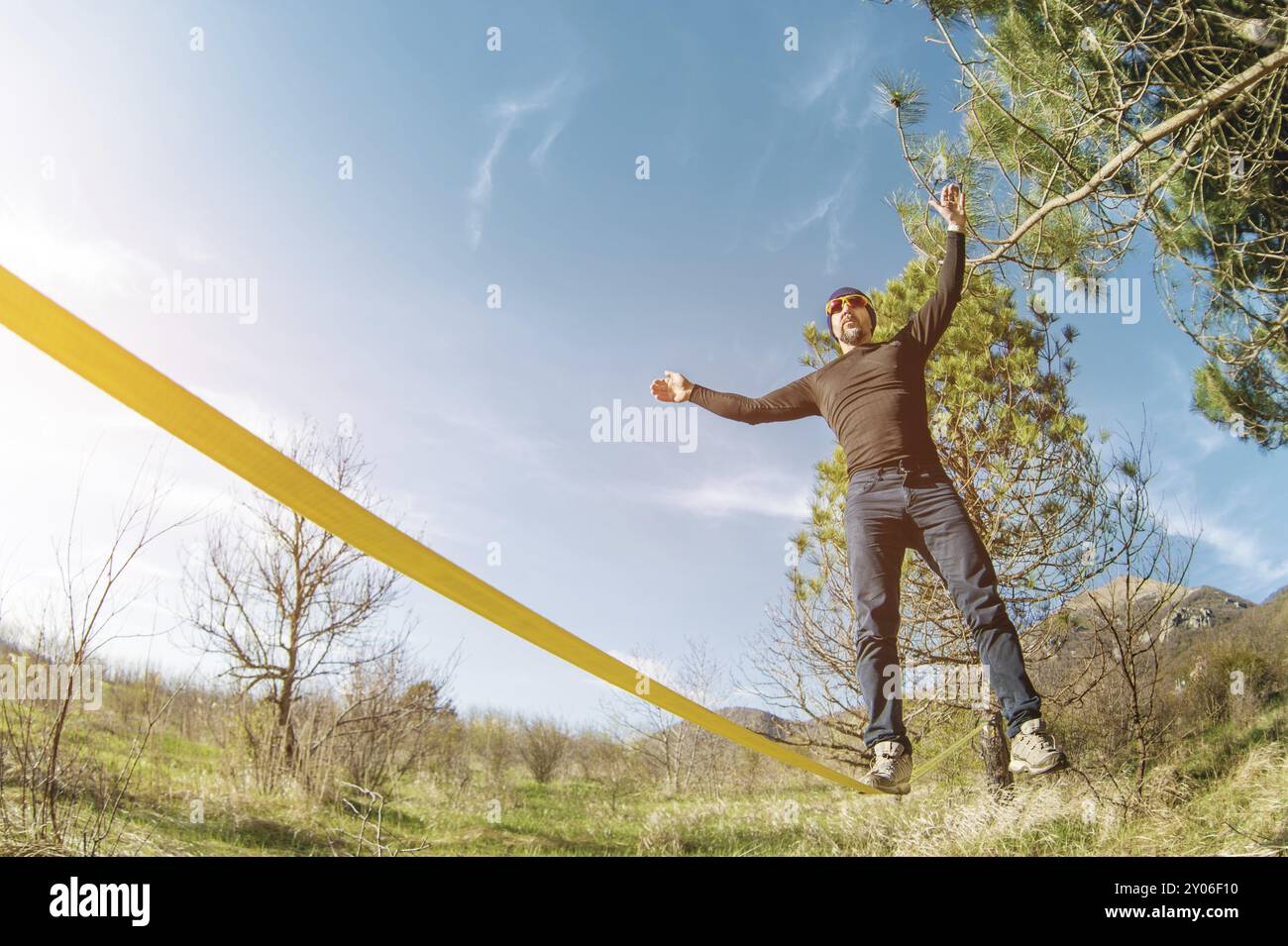 Un uomo, invecchiato con la barba e con gli occhiali da sole, si bilancia su una linea lenta all'aria aperta tra due alberi al tramonto sul cielo blu di sfondo Foto Stock