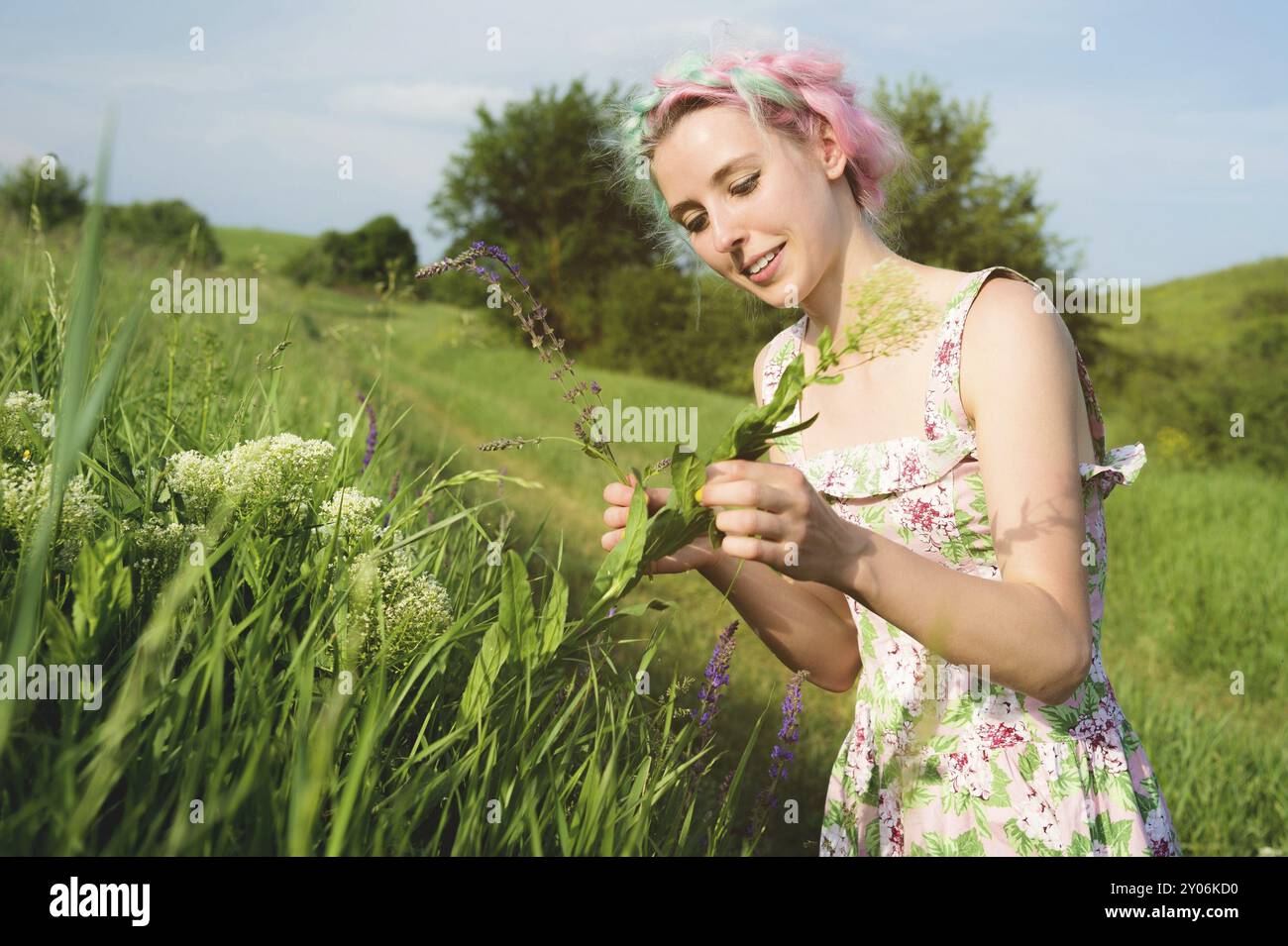 Ritratto di una giovane ragazza felice e carina con capelli multicolori raccoglie fiori accanto a una strada di campagna al tramonto. Il concetto di armonia umana con la n Foto Stock