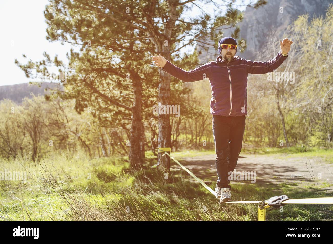 Un uomo, invecchiato con la barba e con gli occhiali da sole, si bilancia su una linea lenta all'aria aperta tra due alberi al tramonto sul cielo blu di sfondo Foto Stock