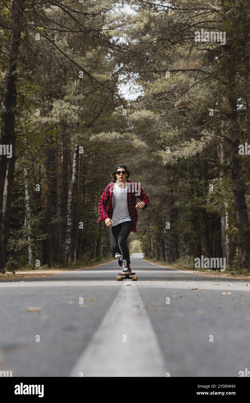 Un giovane hipster con cappello e camicia a quadri e occhiali da sole sta cavalcando il suo skateboard su una strada di campagna in una foresta autunnale. La fase del movimento di d Foto Stock