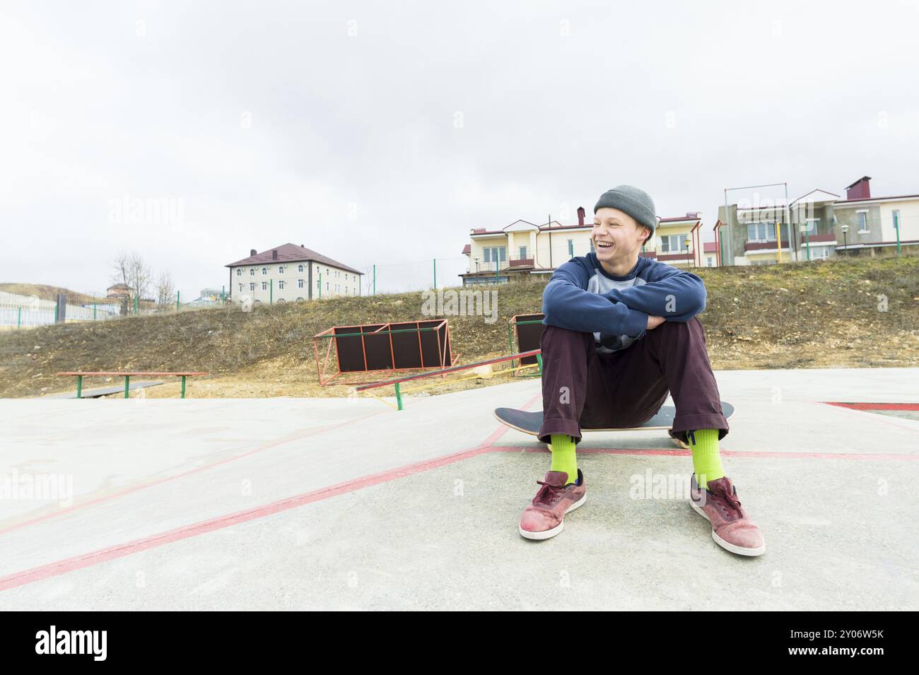 Un ragazzo adolescente è seduto su uno skateboard nel parco e sorridente. Il concetto di passatempo di tempo libero per gli adolescenti in città Foto Stock