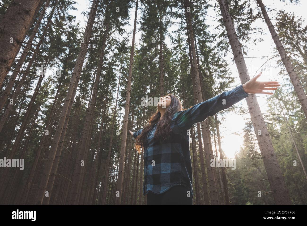 Donna felice in una camicia a quadri con braccia allungate che gode della serenità di una fitta foresta, circondata da alberi alti, sentendo libertà e connessione Foto Stock