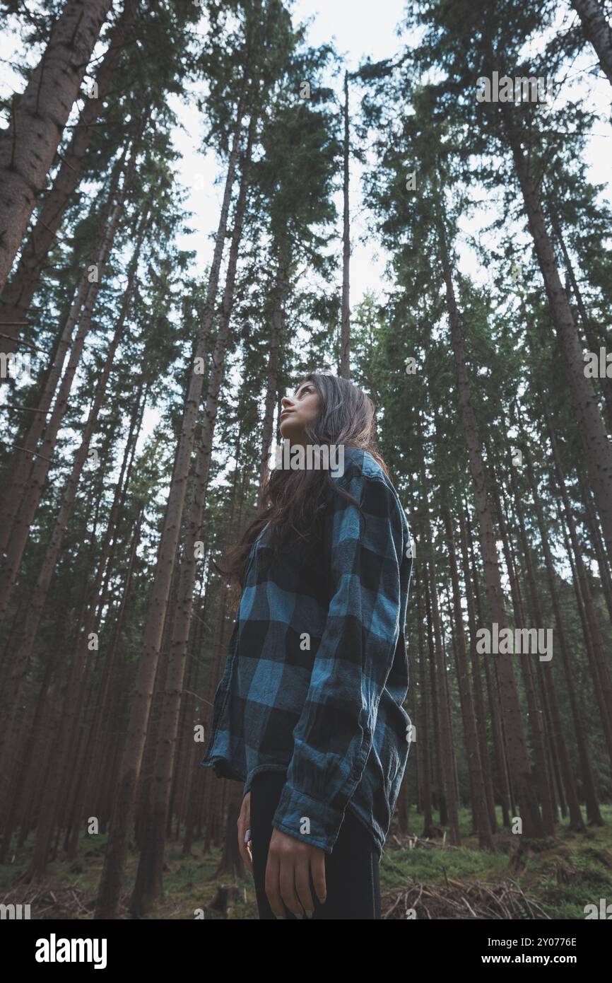 Donna felice in una camicia a quadri che gode della serenità di una fitta foresta, circondata da alberi alti, sentendo libertà e connessione con la natura. Foto Stock
