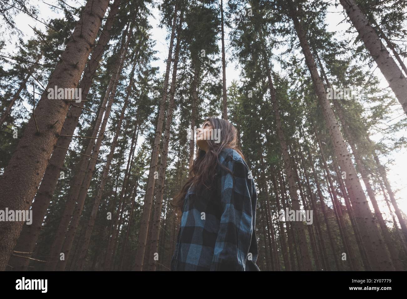Donna felice in una camicia a quadri che gode della serenità di una fitta foresta, circondata da alberi alti, sentendo libertà e connessione con la natura. Foto Stock