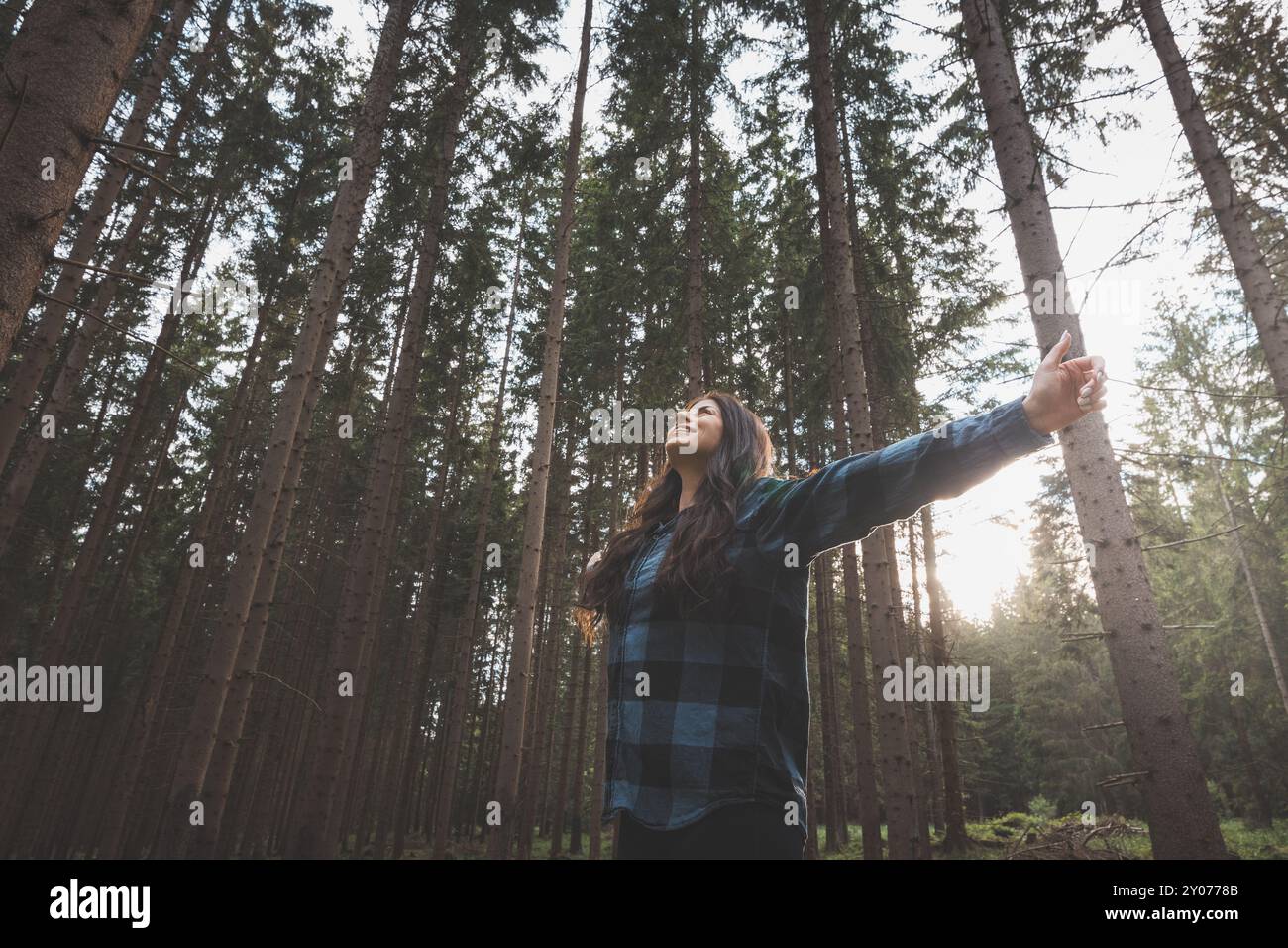 Donna felice in una camicia a quadri con braccia allungate che gode della serenità di una fitta foresta, circondata da alberi alti, sentendo libertà e connessione Foto Stock