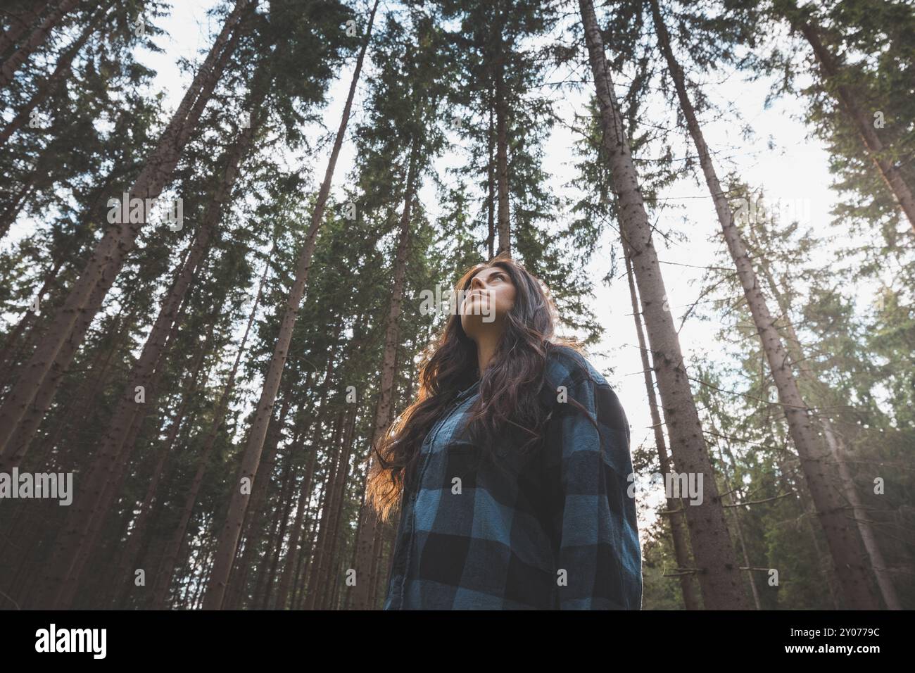 Donna felice in una camicia a quadri con braccia allungate che gode della serenità di una fitta foresta, circondata da alberi alti, sentendo libertà e connessione Foto Stock