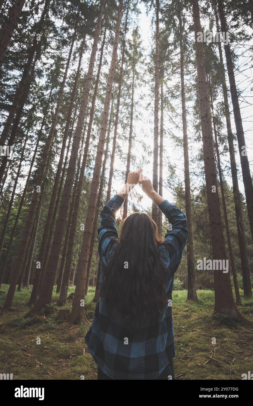 Donna felice in una camicia a quadri che gode della serenità di una fitta foresta, circondata da alberi alti, sentendo libertà e connessione con la natura. Foto Stock