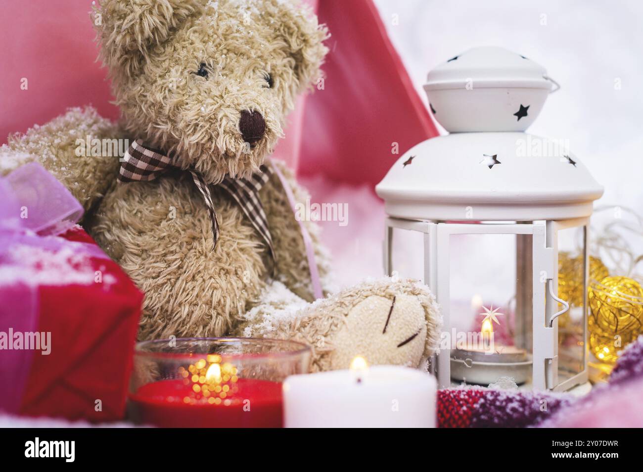 Primo piano di un orsacchiotto con candele di Natale su un accogliente plaid a scacchi all'aperto accanto a una confezione regalo rossa cosparsa di neve. Festività natalizie Foto Stock