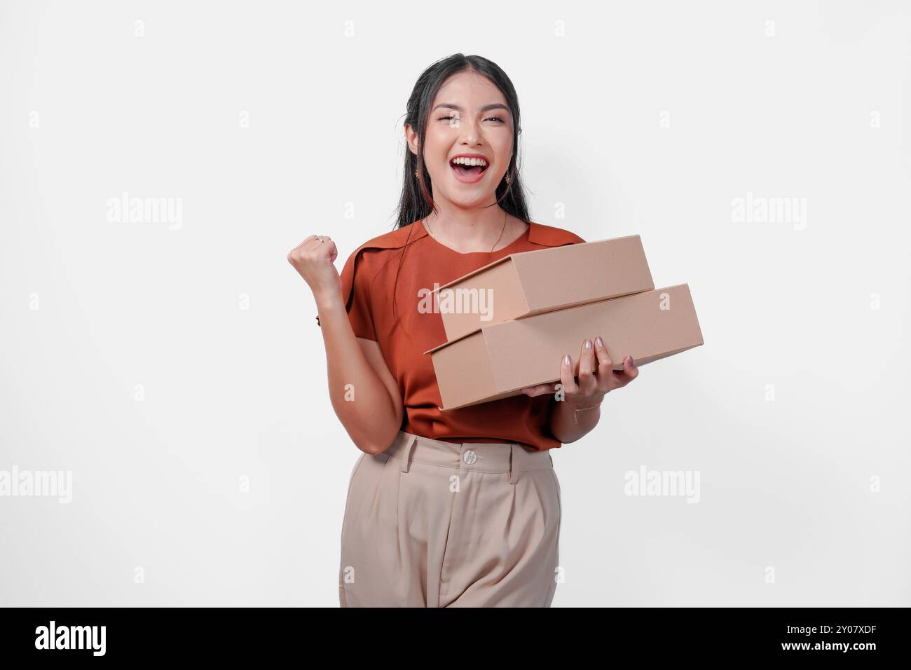 La giovane donna asiatica gioiosa in una camicia marrone regge scatole regalo e solleva le braccia in aria, isolata da sfondo bianco. Concetto di gioia ed exci Foto Stock