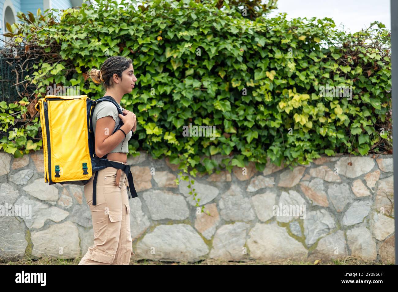 Vista laterale da copyspace di una ragazza ispanica che consegna cibo con uno zaino giallo mentre cammina attraverso un quartiere residenziale per consegnare gli ordini. L'ima Foto Stock