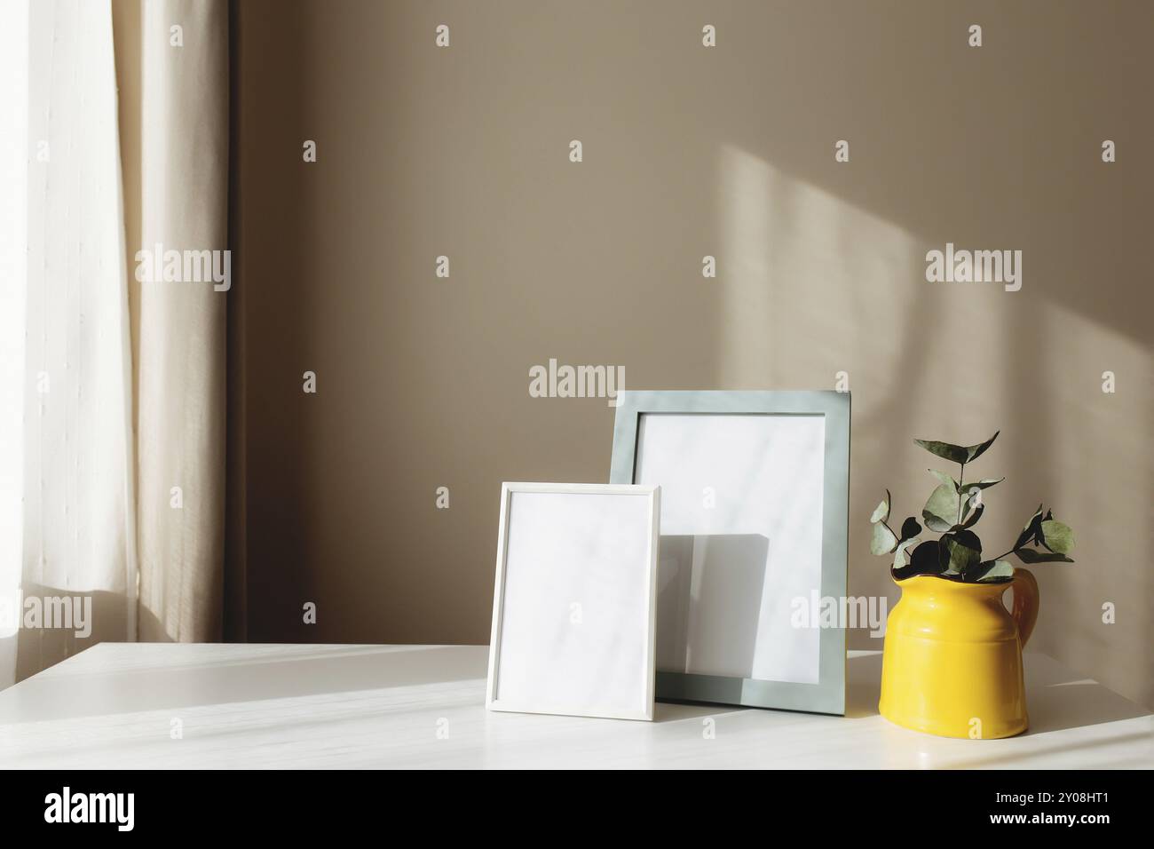 Una caraffa o vaso in ceramica gialla con rami di eucalipto, cornici bianche vuote sul tavolo bianco all'interno con pareti beige vicino alla finestra Foto Stock
