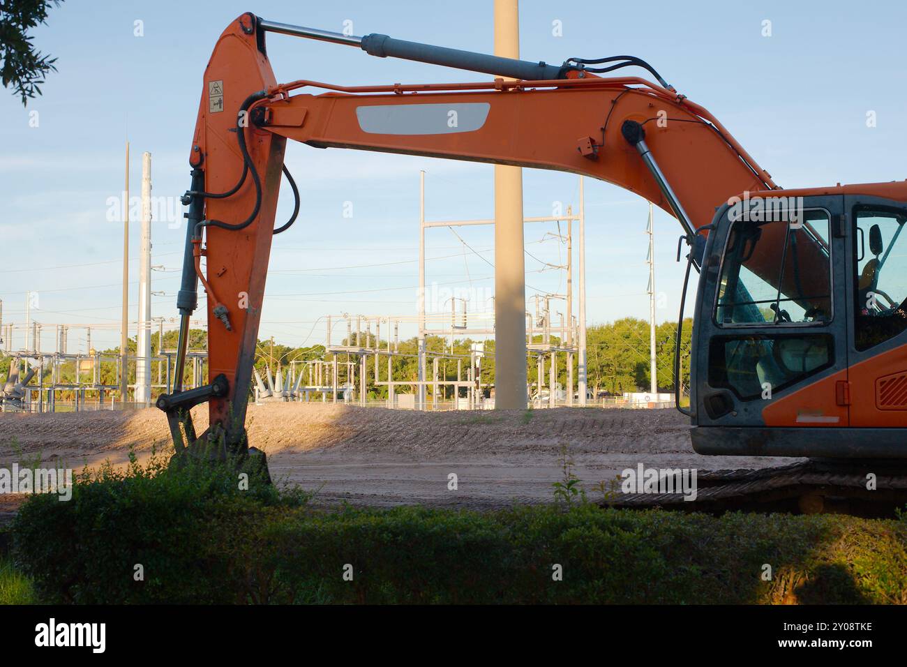 Ampia visuale del telaio attraverso la macchina escavatore a benna arancione nel cantiere della sottostazione elettrica Vista sul cumulo di terra con erba verde davanti Foto Stock