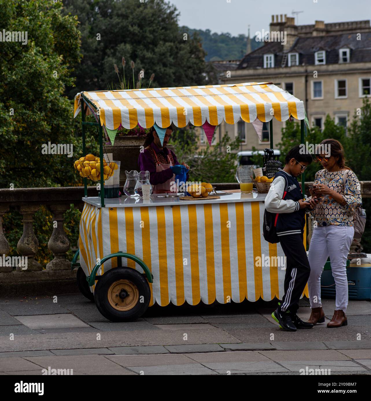 Un piccolo carro di limonate con tendalino a strisce in una pittoresca posizione urbana con due persone nelle vicinanze che discutono qualcosa. Bath, Inghilterra Foto Stock