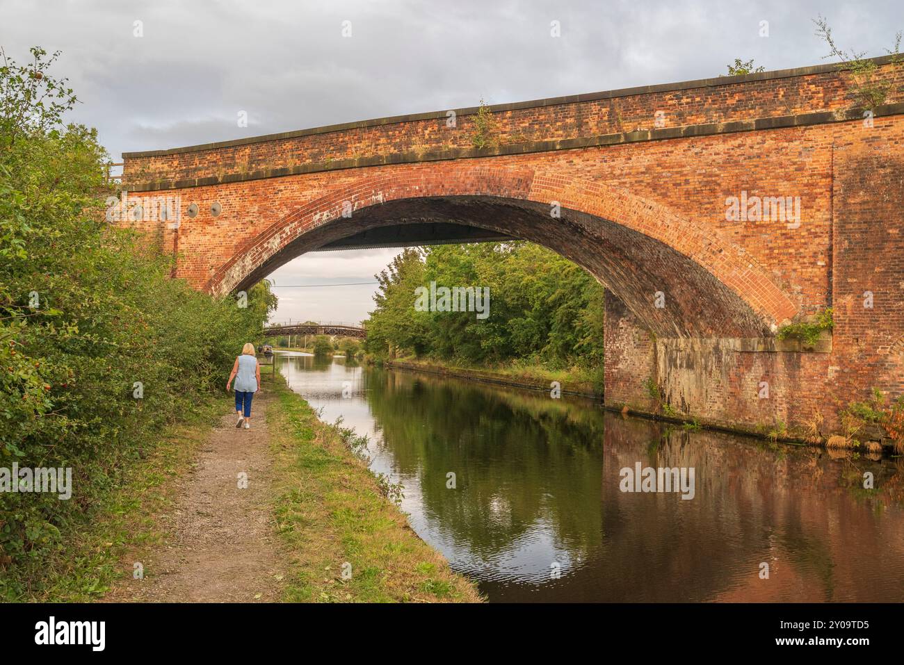 Donna che cammina sul sentiero per le dita dei piedi vicino al canale vicino al ponte ad arco in mattoni nella calda serata estiva, Regno Unito Foto Stock