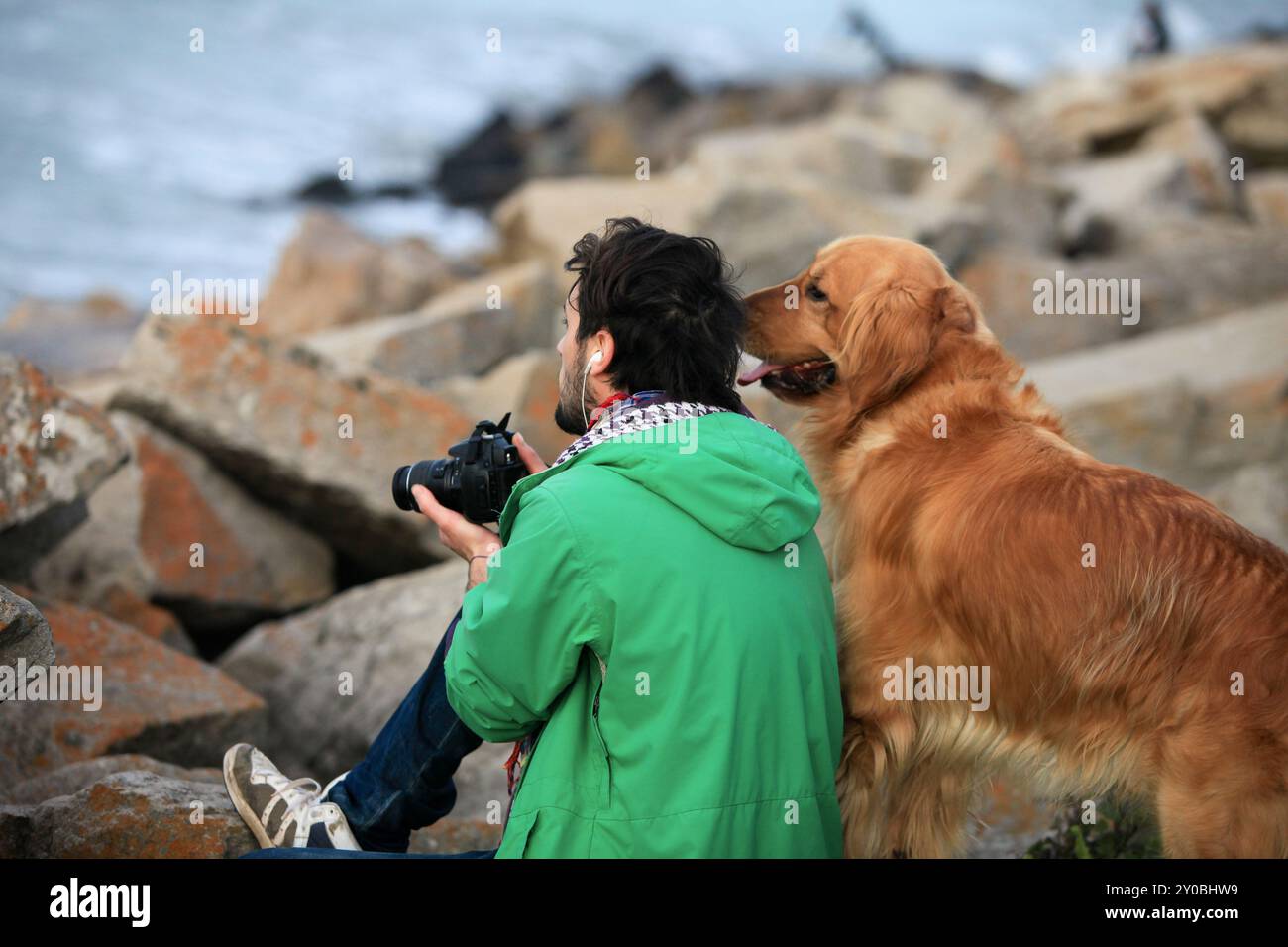 giovane fotografo seduto su rocce costiere con un cane, guardando l'oceano e facendo foto Foto Stock