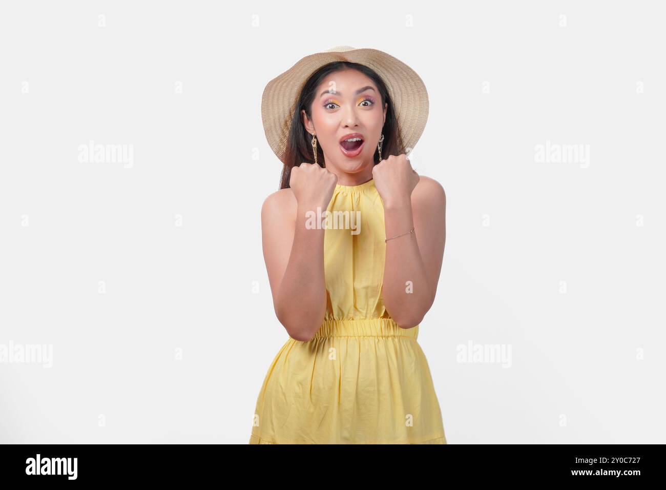 Giovane donna asiatica gioiosa con un vestito giallo che tiene le mani in alto con un'espressione felice di successo. Indossa un cappello di paglia e orecchini Foto Stock