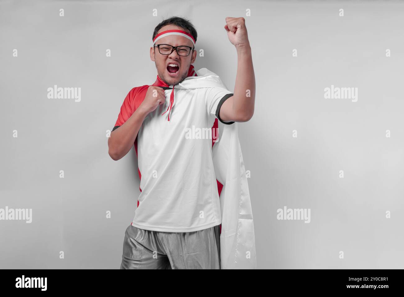 Giovane uomo asiatico energico che indossa una maglia con bandiera drappeggiata sulla spalla alzando la mano in aria con un'espressione felice. Giorno dell'indipendenza dell'Indonesia e. Foto Stock