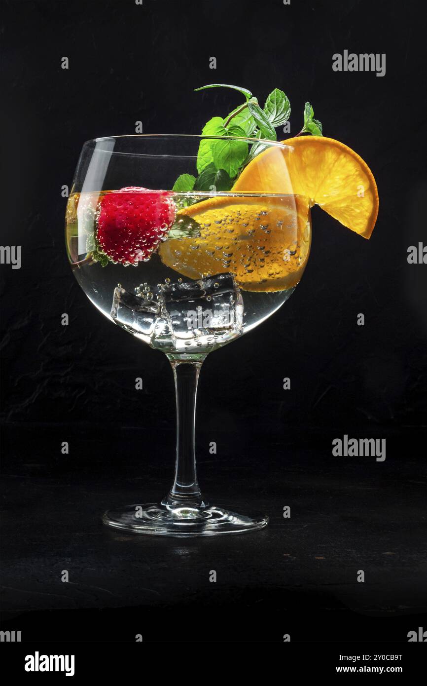 Cocktail elegante con frutta fresca. Gin e bevanda tonica con ghiaccio a una festa, su sfondo nero. Alcol con arancia, menta e fragola Foto Stock
