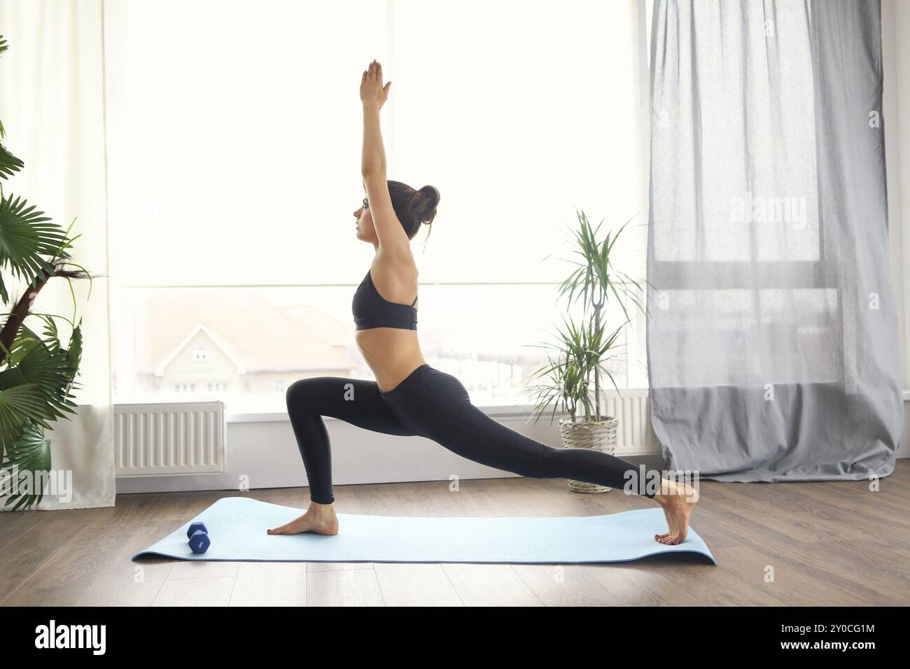 Donna sorridente in sportswear esercitando sul tappetino fitness e guardando la fotocamera in salotto Foto Stock
