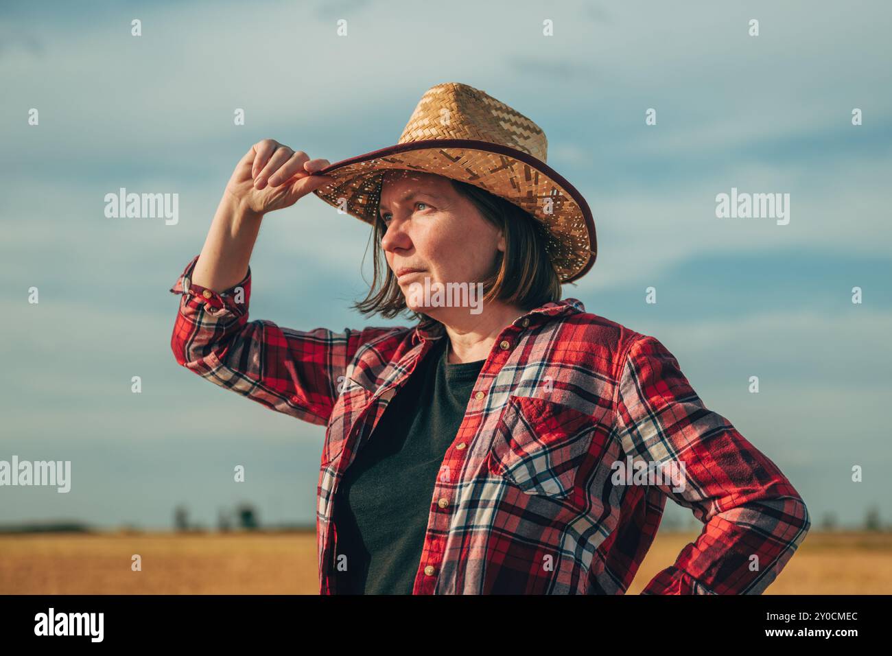 Ritratto di un'agricoltrice che indossa un cappello di paglia in piedi in un campo di grano maturo e che pensa in modo selettivo Foto Stock