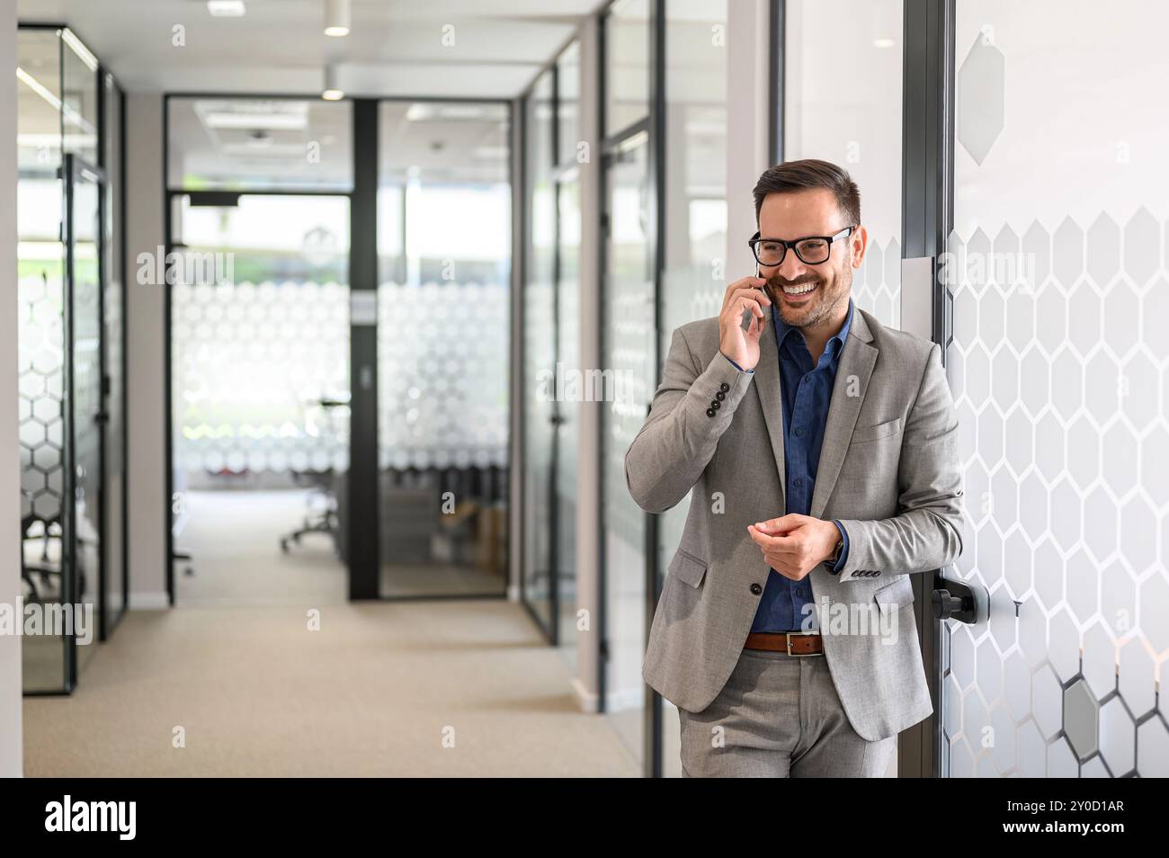 Imprenditore di successo che sorride e parla al telefono cellulare mentre si trova davanti a una porta di vetro in ufficio Foto Stock