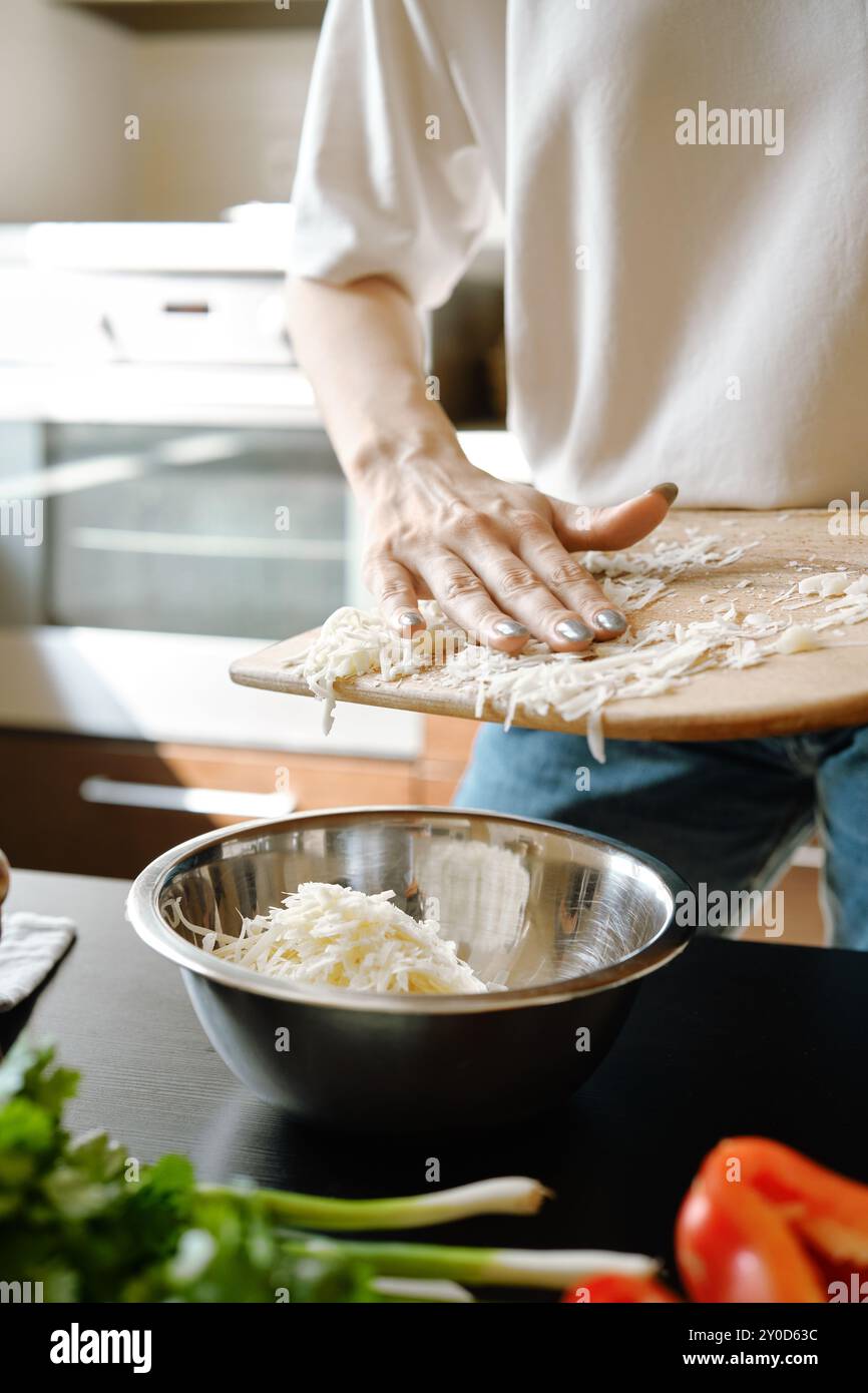 Donna irriconoscibile che versa formaggio grattugiato in una ciotola in cucina Foto Stock