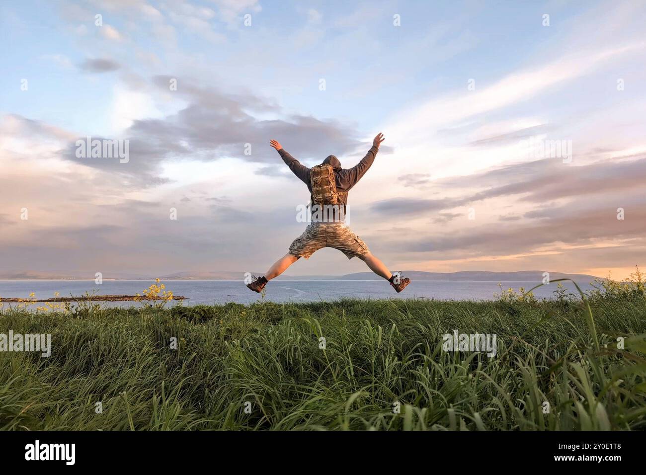 Uomo escursionista in pantaloncini camo e zaino che salta sulla cima della collina verde, mani in alto, vista sulla Wild Atlantic Way a Galway, Irlanda, libertà, avventura e vita Foto Stock