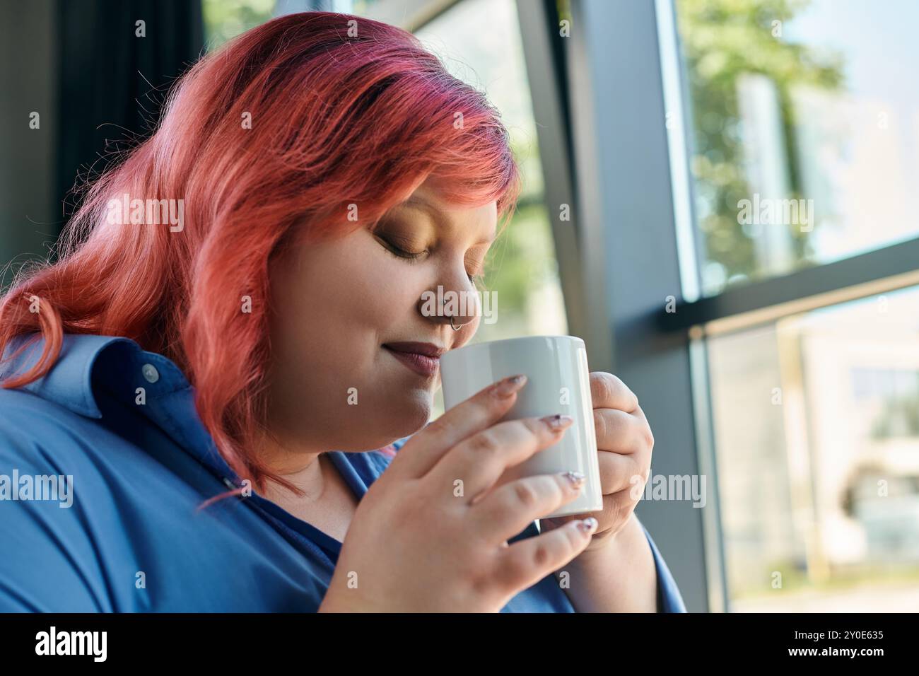 Una donna di grandi dimensioni con i capelli rosa siede accanto a una finestra, gustando una tazza di tè o caffè. Foto Stock