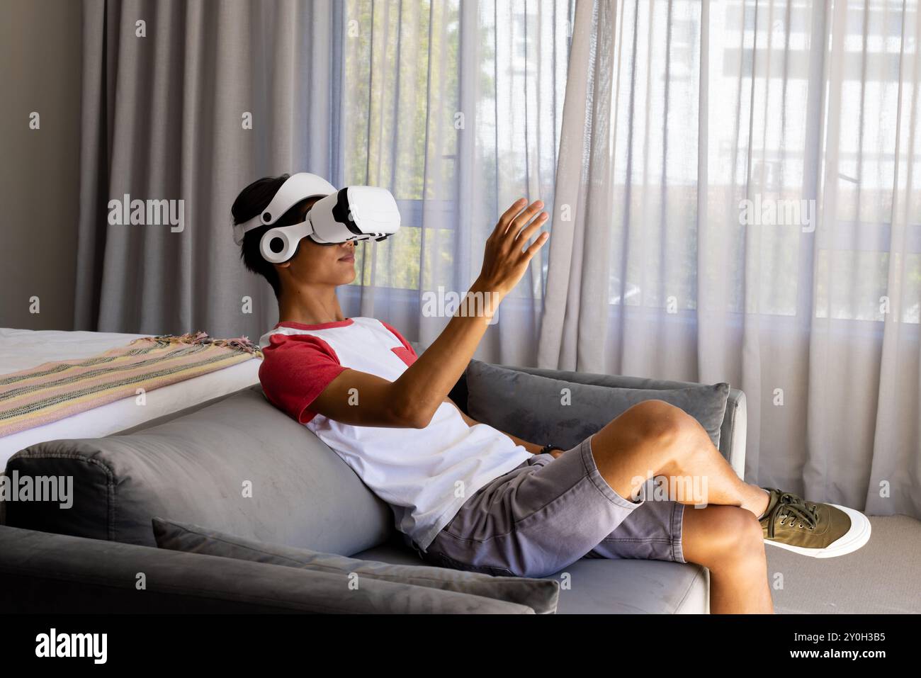 Utilizzando il visore VR, un uomo asiatico seduto sul divano e che interagisce con l'ambiente virtuale Foto Stock