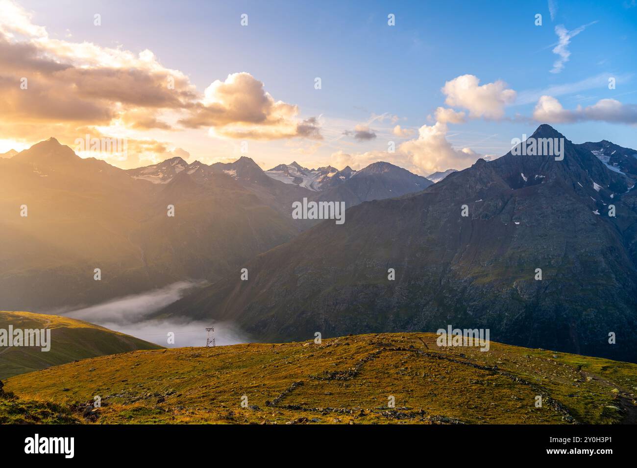 Il sole sorge sulle maestose Alpi Otztal, che proiettano un bagliore dorato sulle vette e sulle valli. Le nuvole si attenuano dolcemente sopra il paesaggio, creando un incredibile contrasto all'alba nella tranquilla regione montana Foto Stock