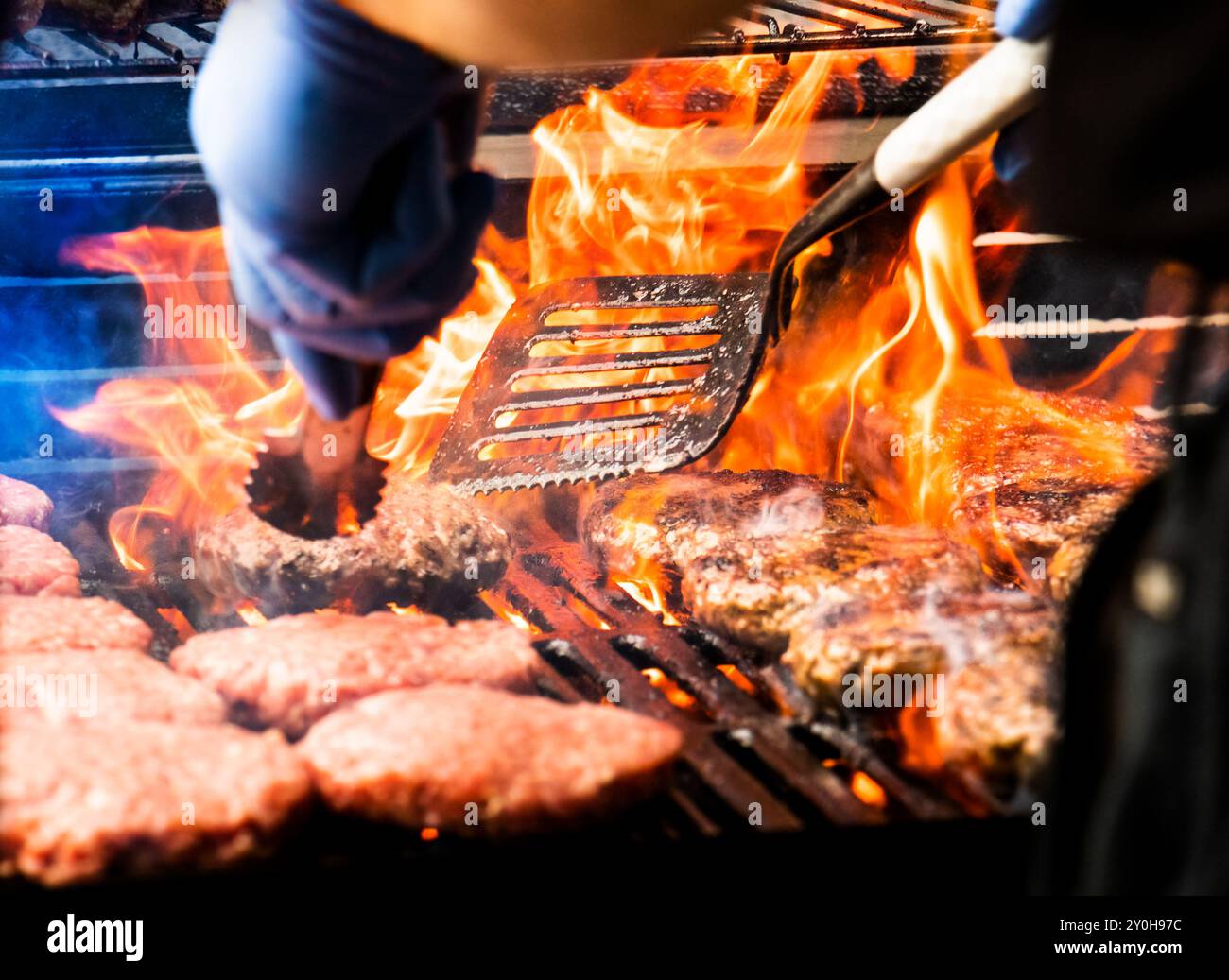 Cottura di hamburger su grill caldo con fiamme - pinze e spatola che muovono polpette grigliate alla fiamma e crude di hamburger alla griglia. Gli hamburger sono tradizionali Foto Stock