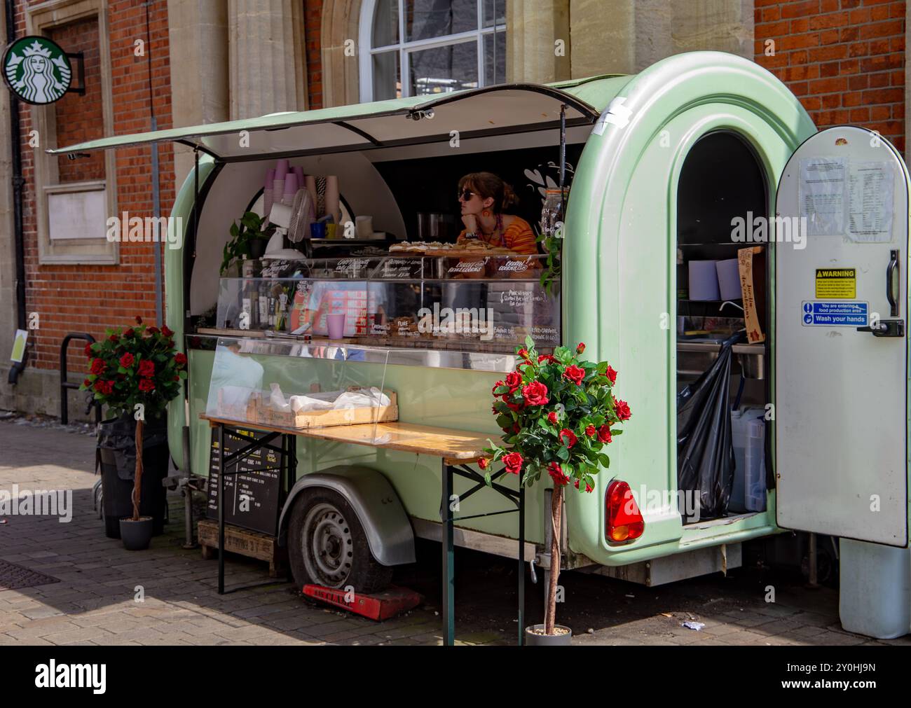 Un food truck in stile vintage che offre una varietà di pasticcini, caffè e bevande, parcheggiato di fronte a un vecchio edificio in mattoni. Foto Stock