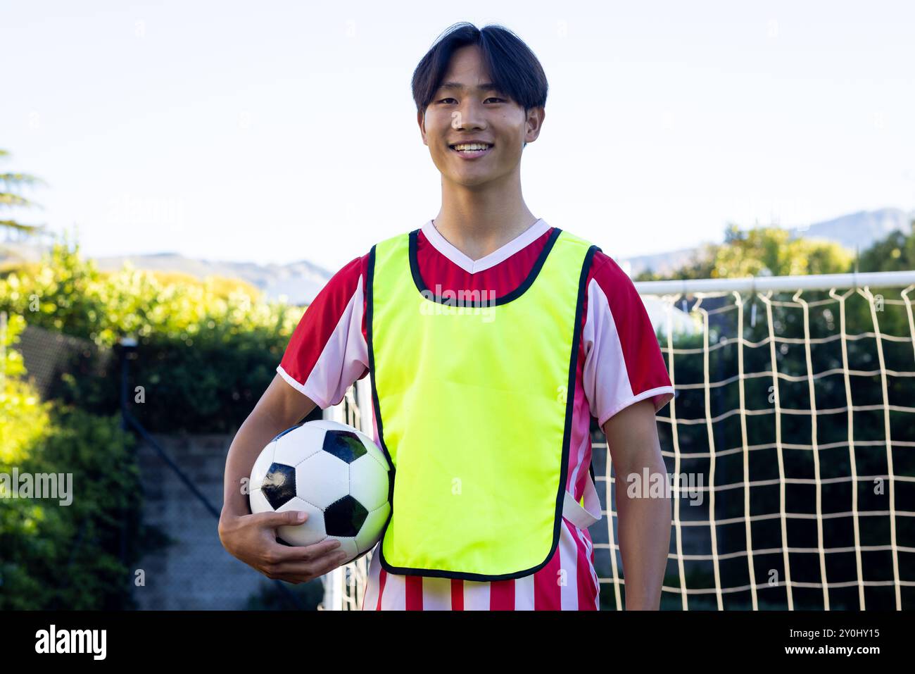 Tenendo palla da calcio, giovane asiatico in uniforme sportiva sorridente per rete porta Foto Stock