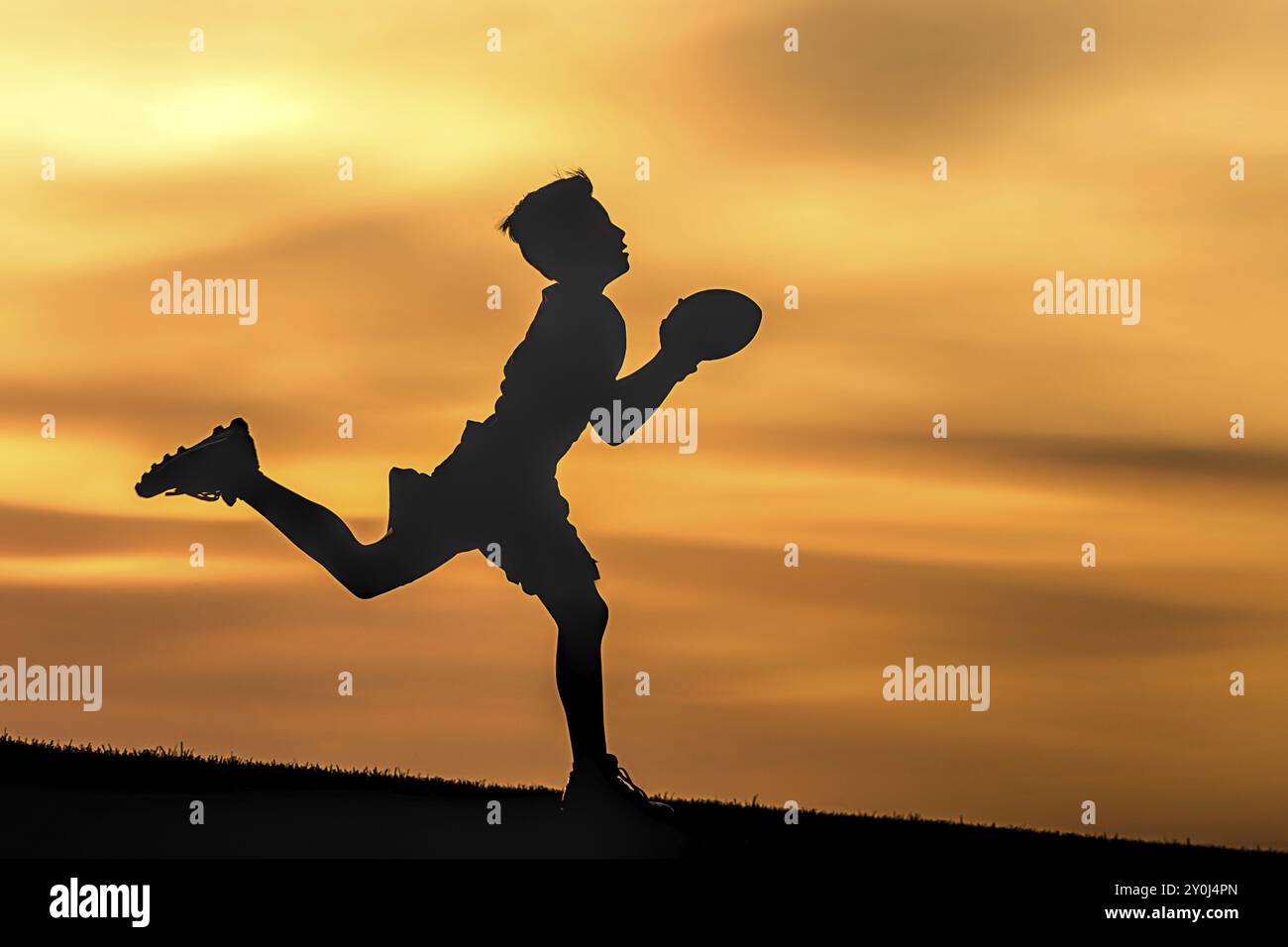 Un ragazzino in silhouette gioca a calcio al tramonto contro un cielo arancione Foto Stock