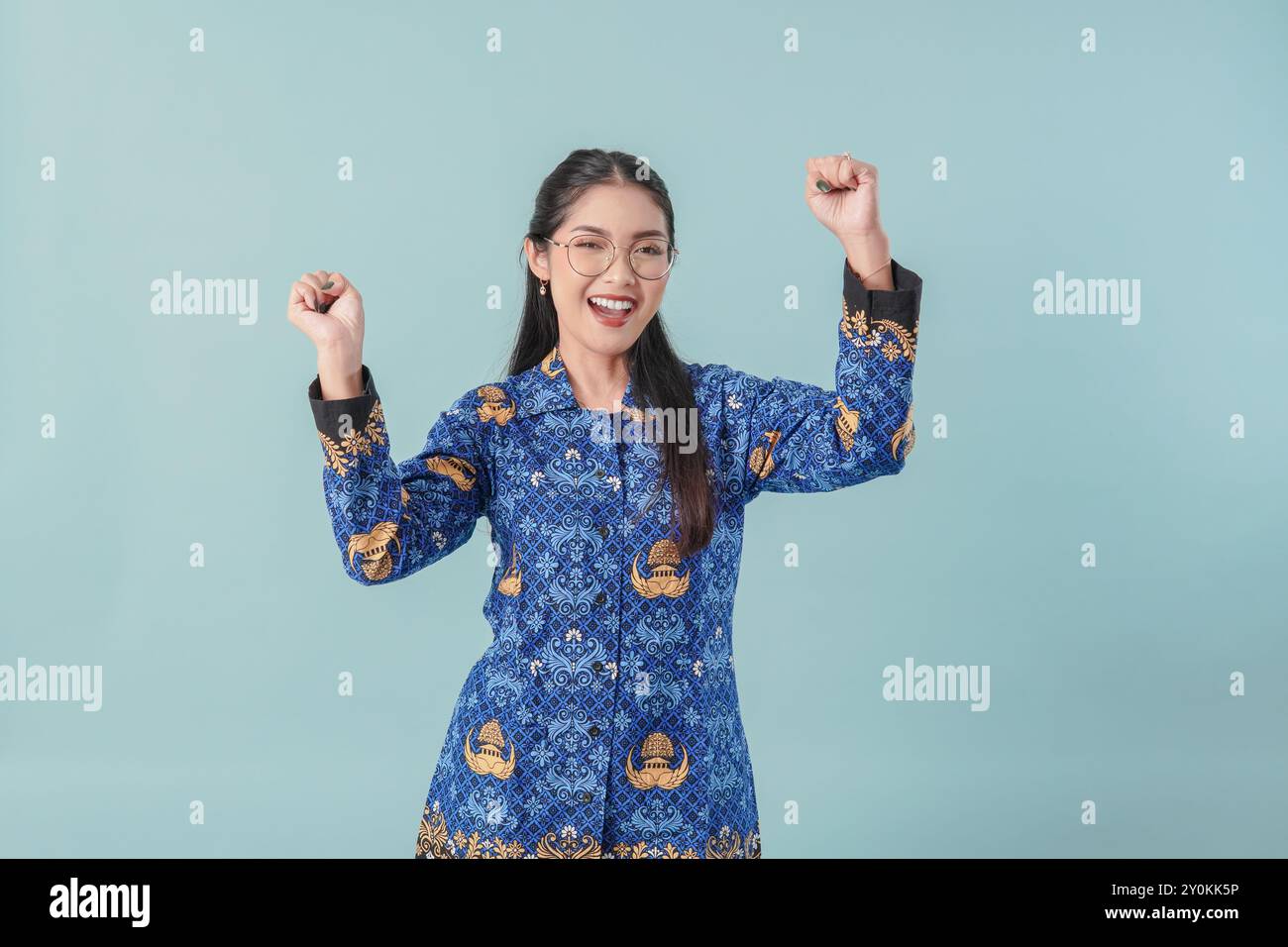 Eccitata donna indonesiana che indossa l'uniforme blu batik e gli occhiali in posa con una felice espressione di successo. Concetto di funzionario pubblico PNS Foto Stock