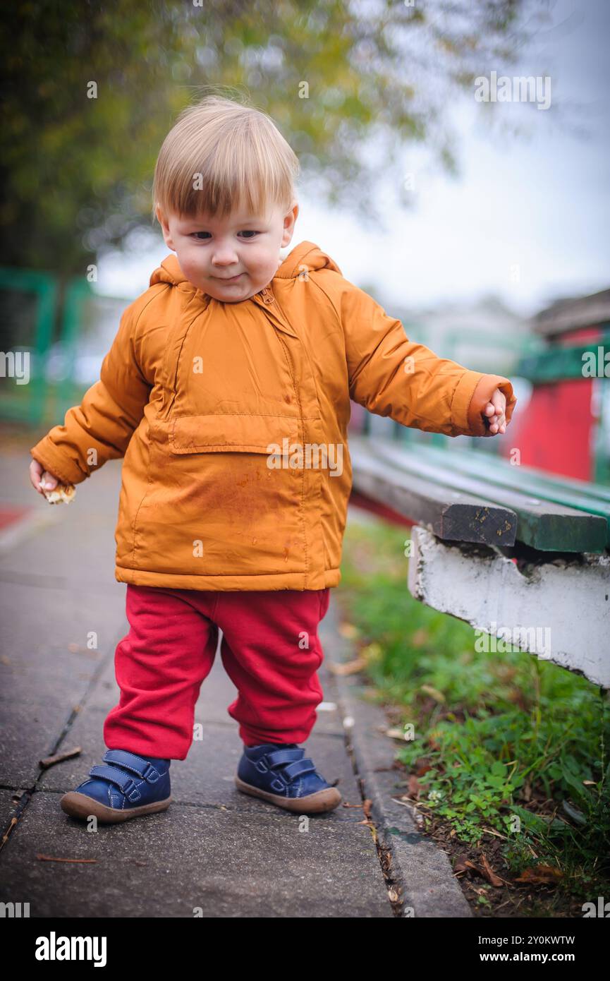 Un bambino allegro compie i suoi primi passi gioiosi lungo un percorso per il parco giochi, racchiuso in una calda giacca arancione e pantaloni rossi, assaporando l'aria frizzante dell'autunno Foto Stock