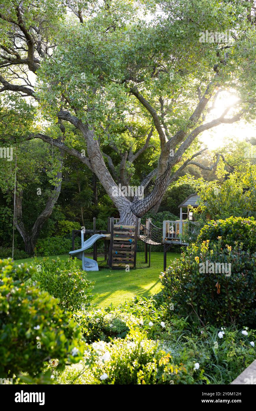 Casa sull'albero e parco giochi nel lussureggiante giardino con luce solare che filtra tra gli alberi Foto Stock