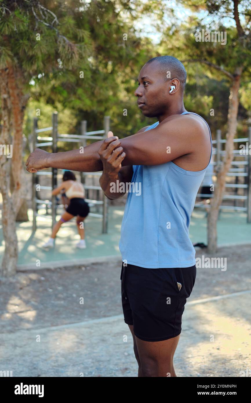 Atletico africano vestito con camicia blu senza maniche, pantaloncini neri fanno stretching delle spalle e scaldano gli esercizi nel parco, nella soleggiata giornata estiva. Sport, guarisci Foto Stock