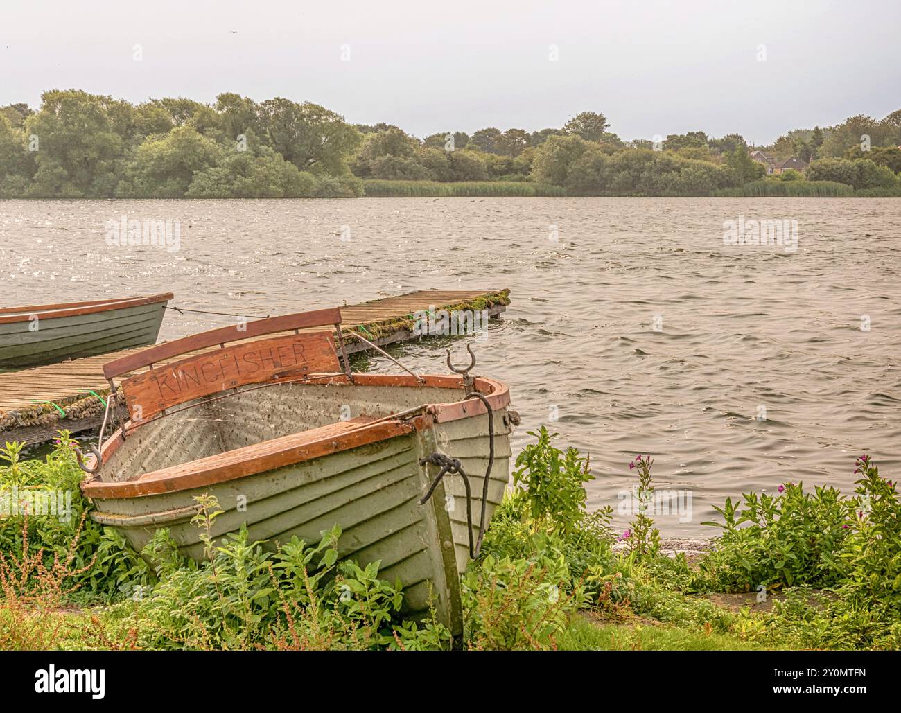 Una barca a remi si arena sulla riva di un lago. Un molo finisce nell'acqua e c'è un bosco sulla riva lontana. Sopra c'è un cielo limpido. Foto Stock