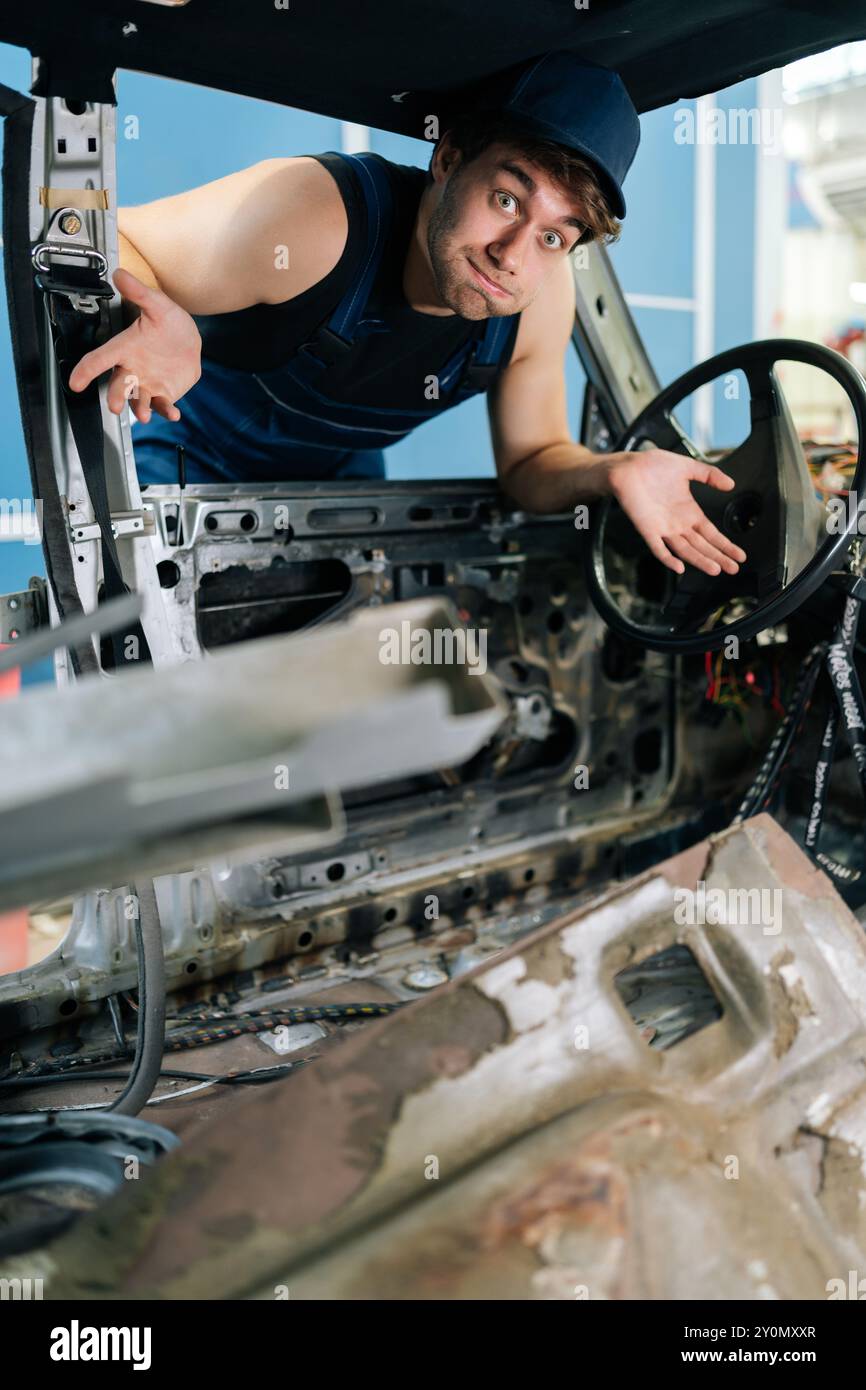 Ritratto verticale del movimento maschile meccanico sconcertato all'interno di un'auto smontata, che mostra l'esperienza nel settore automobilistico e i lavori in corso, guardando la fotocamera Foto Stock