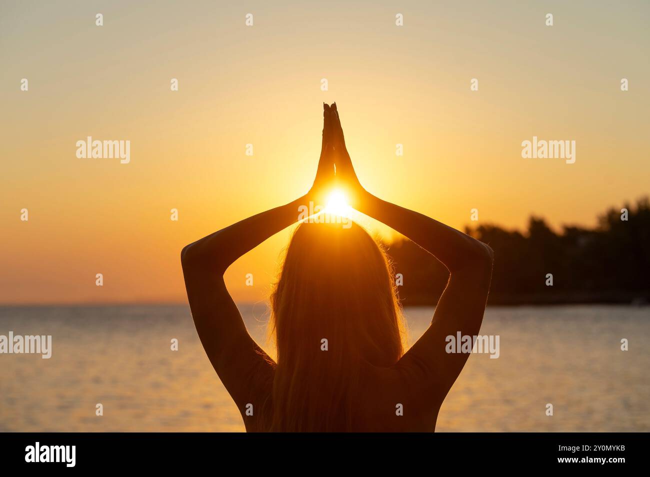 Vista da dietro di una donna con lunghi capelli in piedi in riva al mare che pratica consapevolezza e gratitudine. Tenendole il palmo Unito sopra la testa con il sole Foto Stock