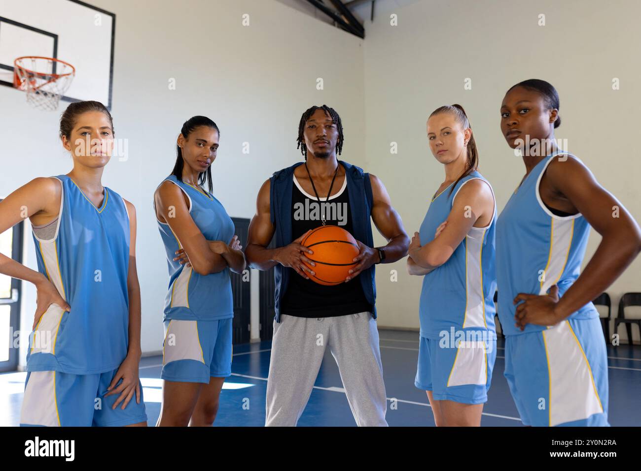 Allenatore afroamericano di basket maschile che tiene la palla con una squadra femminile in palestra, posa con fiducia Foto Stock