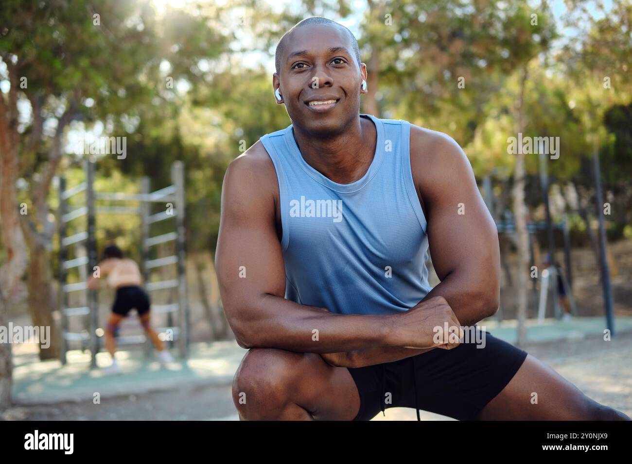 Atletico africano vestito con camicia blu senza maniche, pantaloncini neri allungano le gambe o riscaldano l'esercizio fisico nel parco pubblico nella soleggiata giornata estiva. Sport Foto Stock